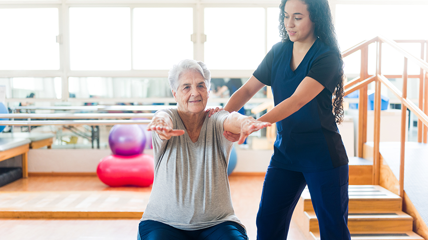 Elderly woman using balance ball helped by young woman in medical scrubs.