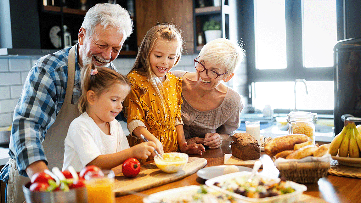 Photograph of a family gathered around a kitchen table preparing and enjoying a meal together. The table features various foods including bread, salad, fruit, and juice, highlighting a shared, casual dining experience in a bright, modern kitchen.