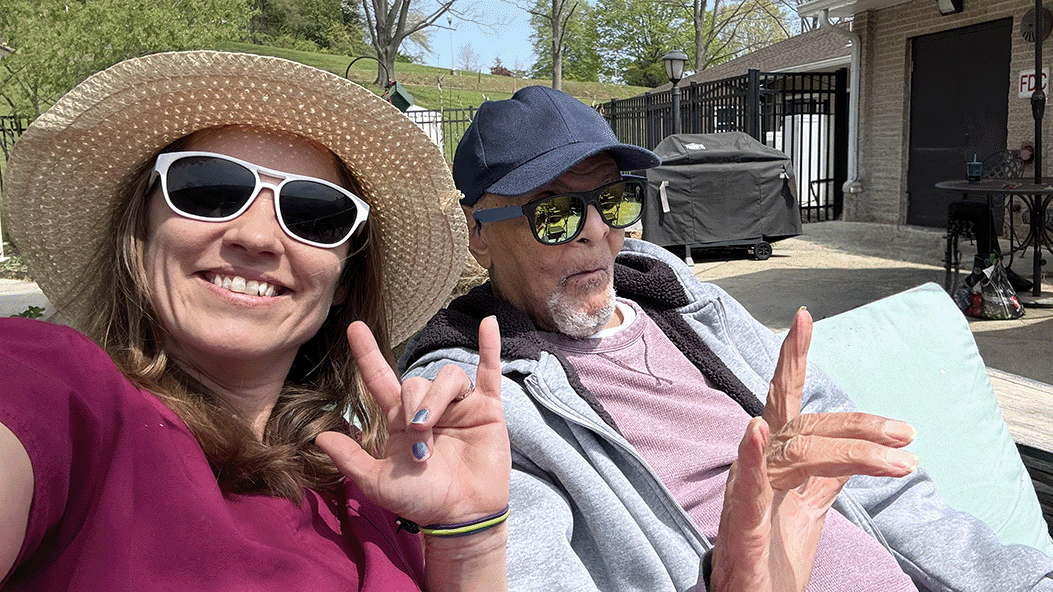 Photograph of two people sitting outdoors on a couch, making hand signs with their fingers. One person wears a large straw hat and a purple shirt, while the other wears a blue cap and a gray hoodie, with a grill and patio area visible in the background.