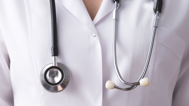 Photograph showing a close-up of a person wearing a white medical coat with a stethoscope draped around their neck. The focus is on the stethoscope's chest piece and tubing, highlighting medical profession context.