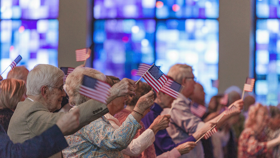 Photograph of a group of elderly people seated indoors, waving small American flags during a patriotic event. The scene features stained glass windows in the background, with attendees focused on the celebration, highlighting a sense of community and national pride.