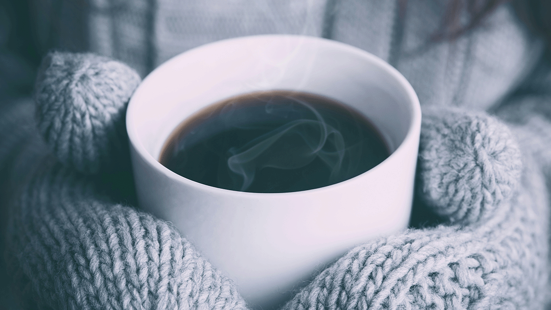 Photograph of a white ceramic cup filled with steaming hot coffee held by hands wearing thick, gray knitted mittens. The background is blurred, emphasizing warmth and coziness in a cold outdoor setting.