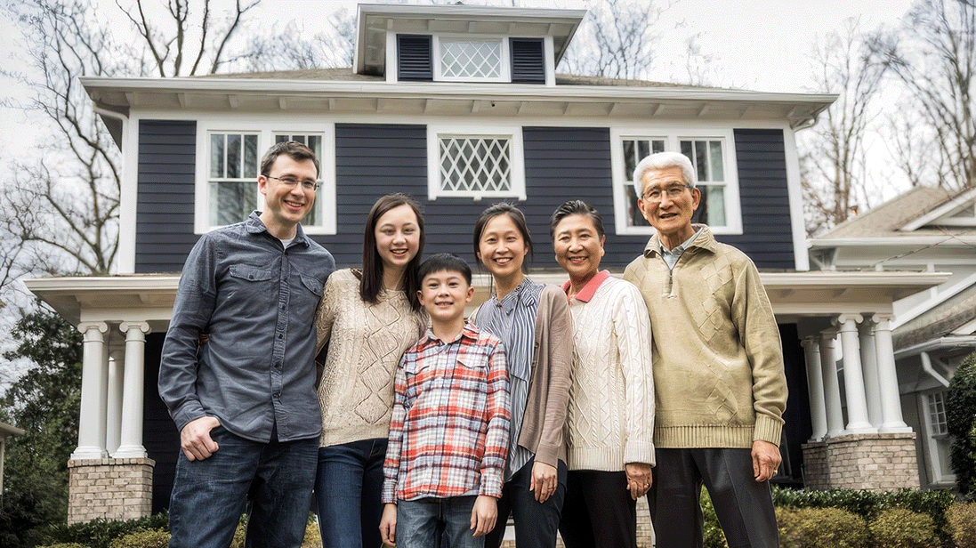 Photograph shows six people, including a child, posing together outside a dark-painted house with white columns, trim, and dormer windows. Group appears to be a multigenerational family portrait, with leafless trees and neighboring homes visible in background.