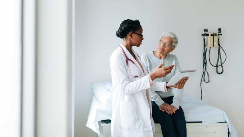 Image shows a healthcare consultation, with a physician in a white coat discussing information on a tablet with an older patient seated on an examination bed. Bright clinical room includes medical equipment, wall-mounted diagnostic devices, and a calm, professional atmosphere.